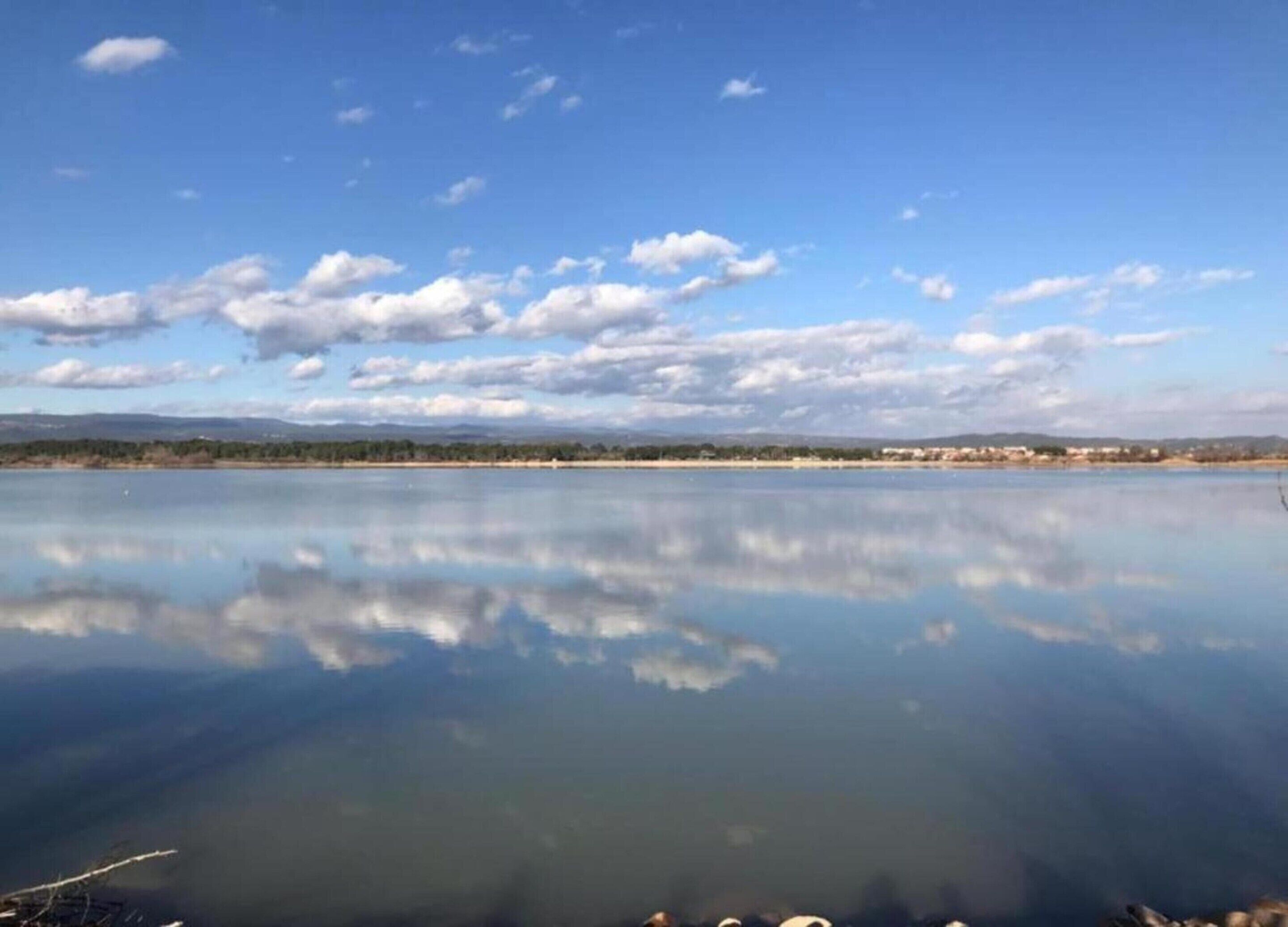 A calm lake with a clear reflection of the sky and clouds, surrounded by a flat landscape with distant buildings.