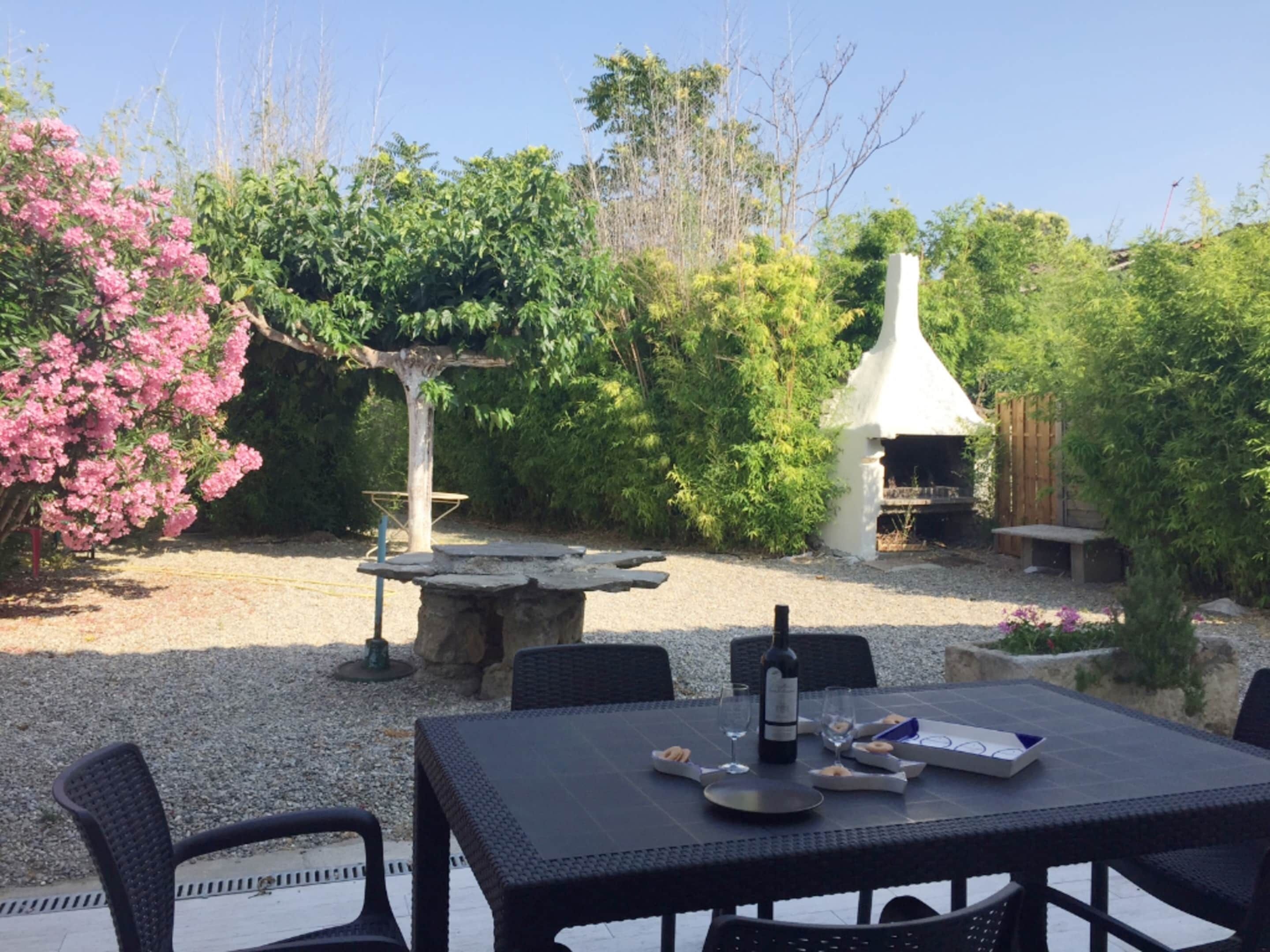 An outdoor patio with a table set for dining, surrounded by greenery and a white structure.
