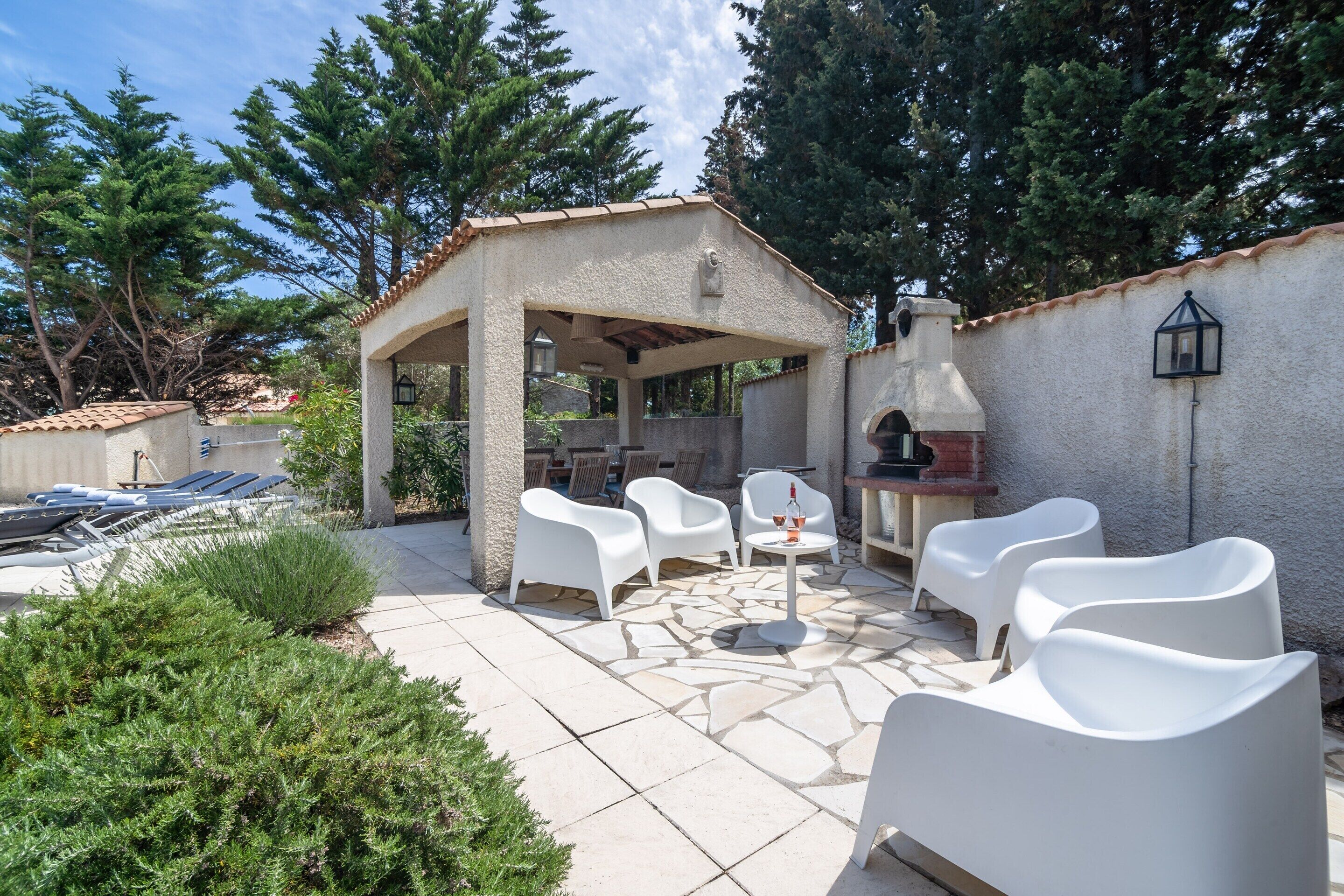 An outdoor patio with white chairs and a small table, surrounded by greenery and a stone-tiled floor.