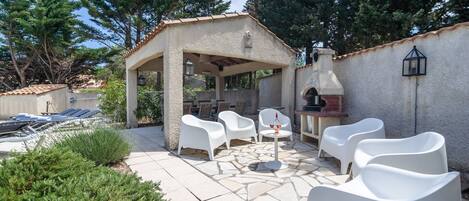 An outdoor patio with white chairs and a small table, surrounded by greenery and a stone-tiled floor.
