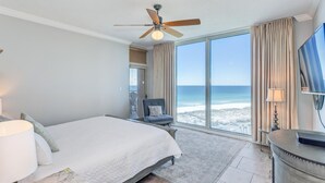 Hotelkamer met een bed, een stoel, een bureau, een plafondventilator en een groot raam met uitzicht op het strand.