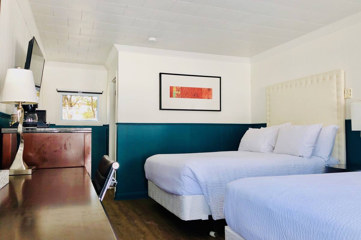 Quarto de hotel com duas camas, uma escrivaninha com luminária, uma cafeteira e um quadro na parede.