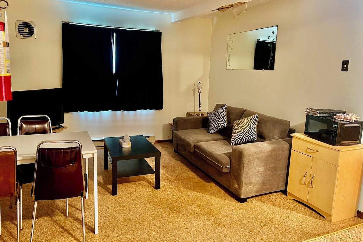 Una sala de estar con un sofá, una mesa de centro, un televisor, una mesa de comedor con sillas y un microondas sobre un mueble.