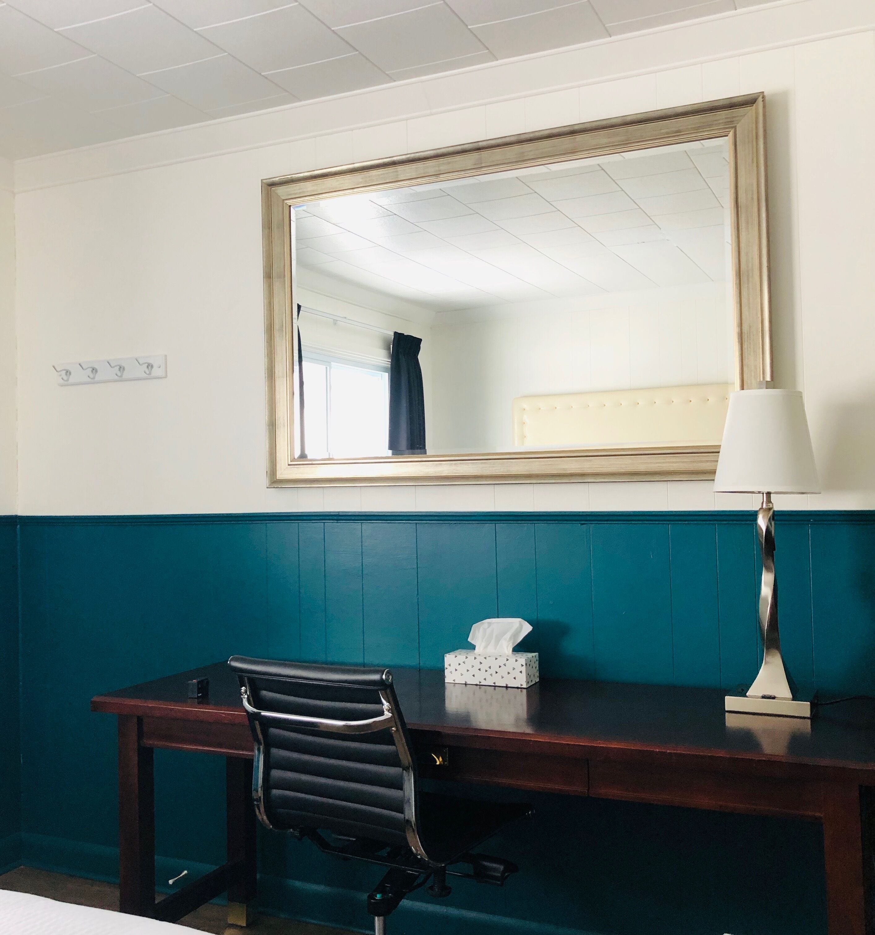A hotel room with a wooden desk, a black office chair, a bedside lamp, and a large mirror on the wall.