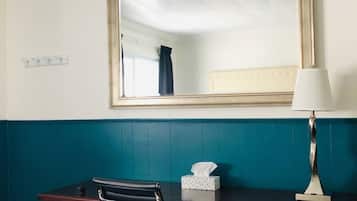 A hotel room with a wooden desk, a black office chair, a bedside lamp, and a large mirror on the wall.