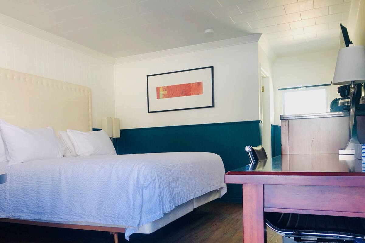 Quarto de hotel com cama, escrivaninha, cadeira, abajur e quadro na parede.
