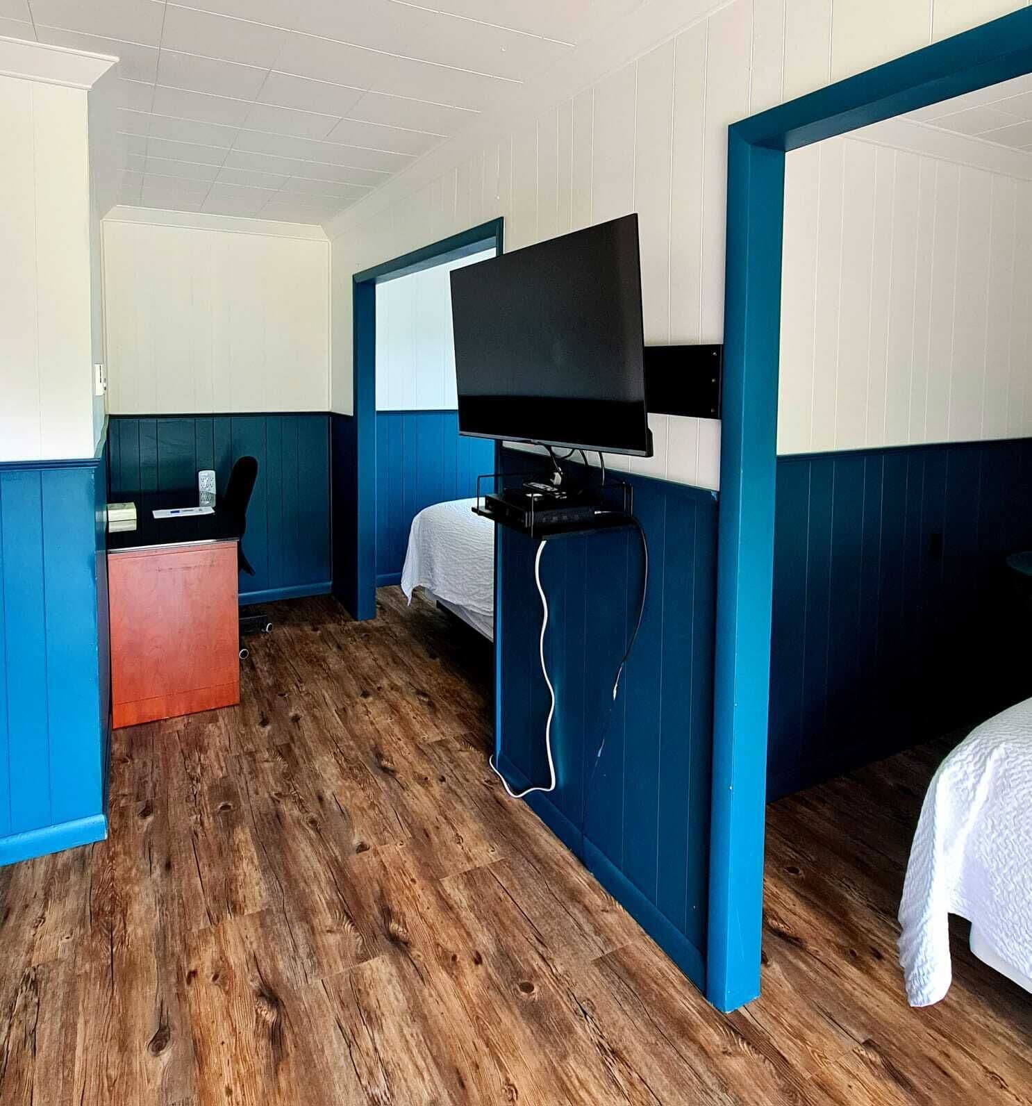 A room with a wooden floor, a bed, a desk with a chair, and a television mounted on the wall.