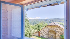 A window with a view of a stone building and a landscape with trees and hills. - Vic-sur-cere Family Nest (Vic-sur-Cere)