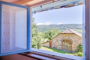 A window with a view of a stone building and a landscape with trees and hills. - Vic-sur-cere Family Nest (Vic-sur-Cere)