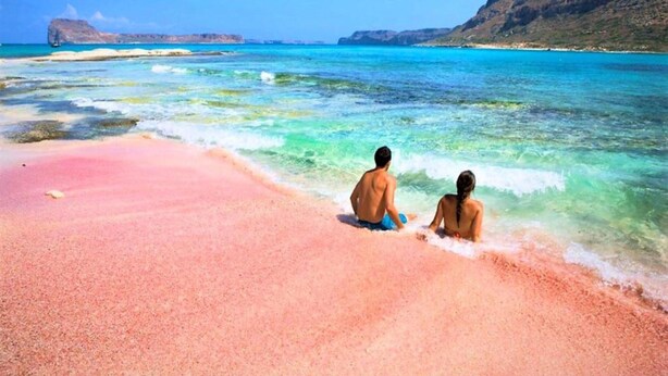 Ein Strand mit klarem türkisblauem Wasser, einem rosafarbenen Sandstrand und in der Ferne felsigen Klippen.
