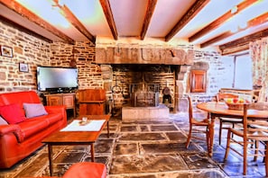 A stone-floored room with a fireplace, a red sofa, a wooden table, and a television. - Alluring Holiday Home With Fenced Garden, Barbecue (Montaigu-les-Bois)
