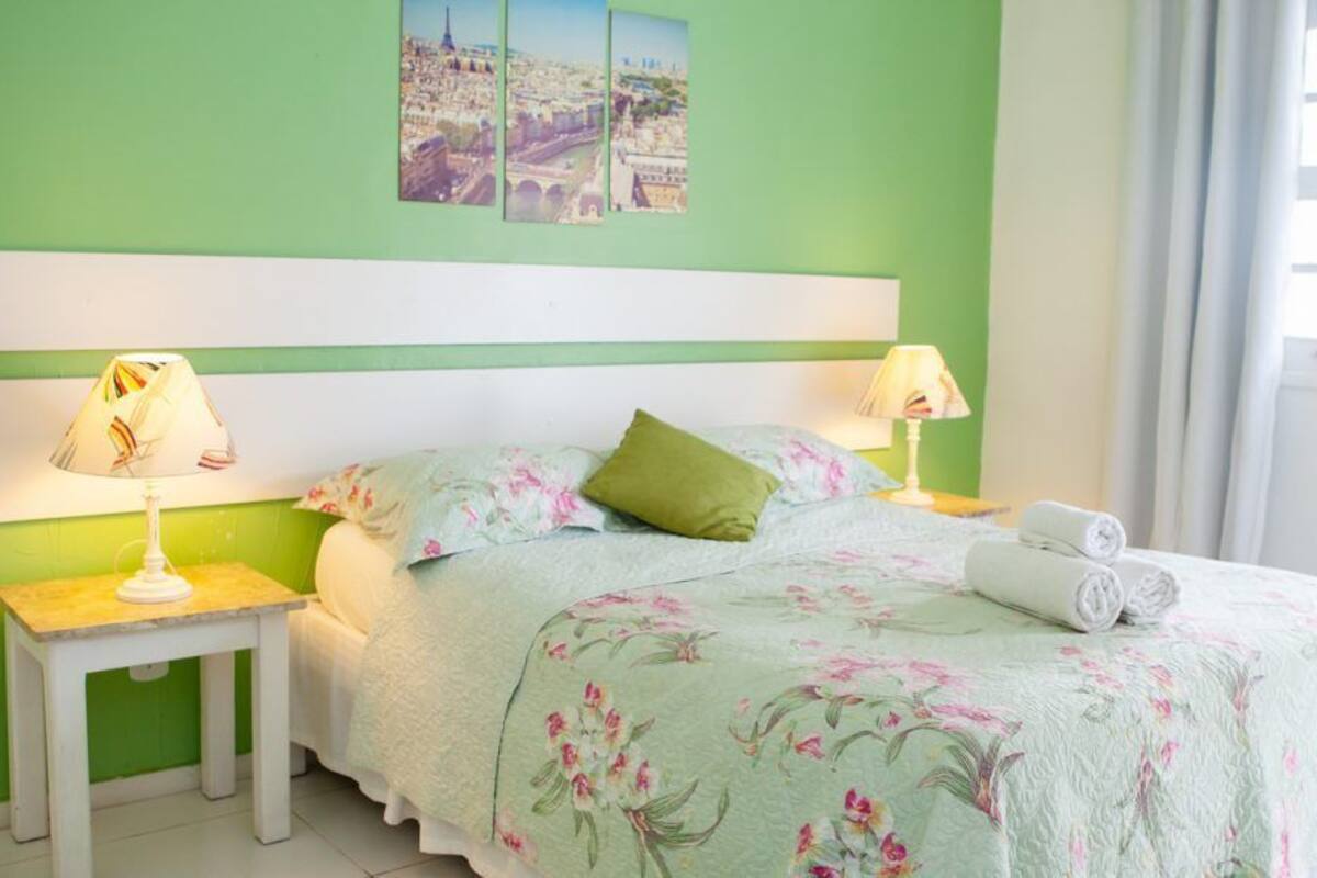 Una cama con ropa de cama floral, dos lámparas de noche, un cabecero verde y una ventana con cortinas.
