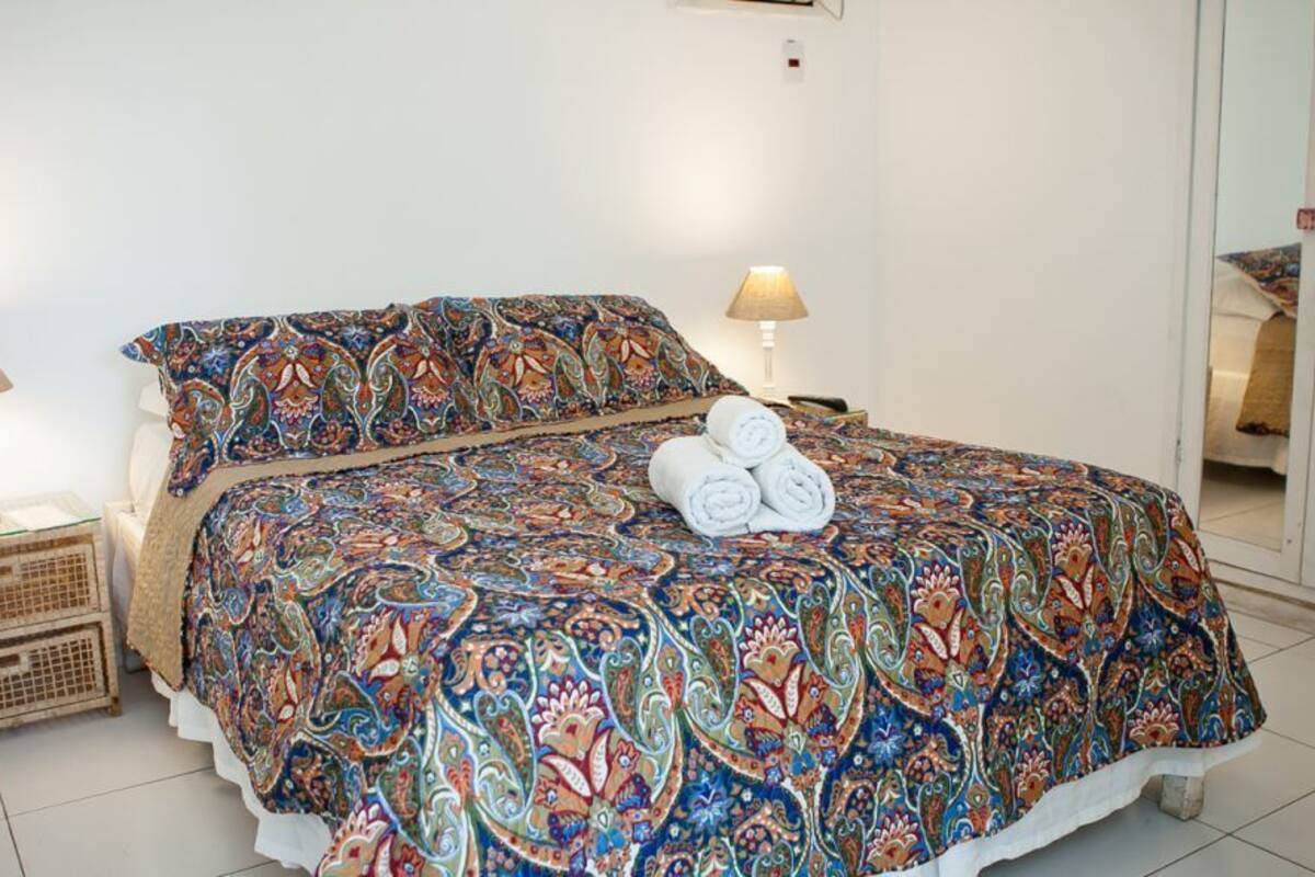 Una cama bien hecha con una colcha estampada, dos lámparas de noche y una pequeña cesta de mimbre sobre un suelo de baldosas.