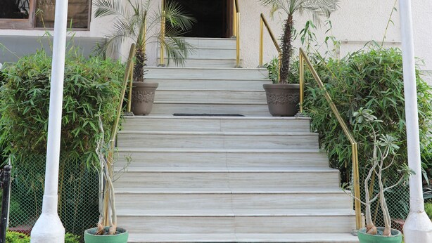 Una escalera conduce a la entrada de un edificio, con un porche cubierto, plantas en macetas y un felpudo.