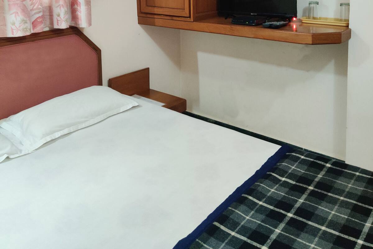 A bedroom with a bed, a wooden wardrobe, a television on a shelf, and a window with floral curtains.