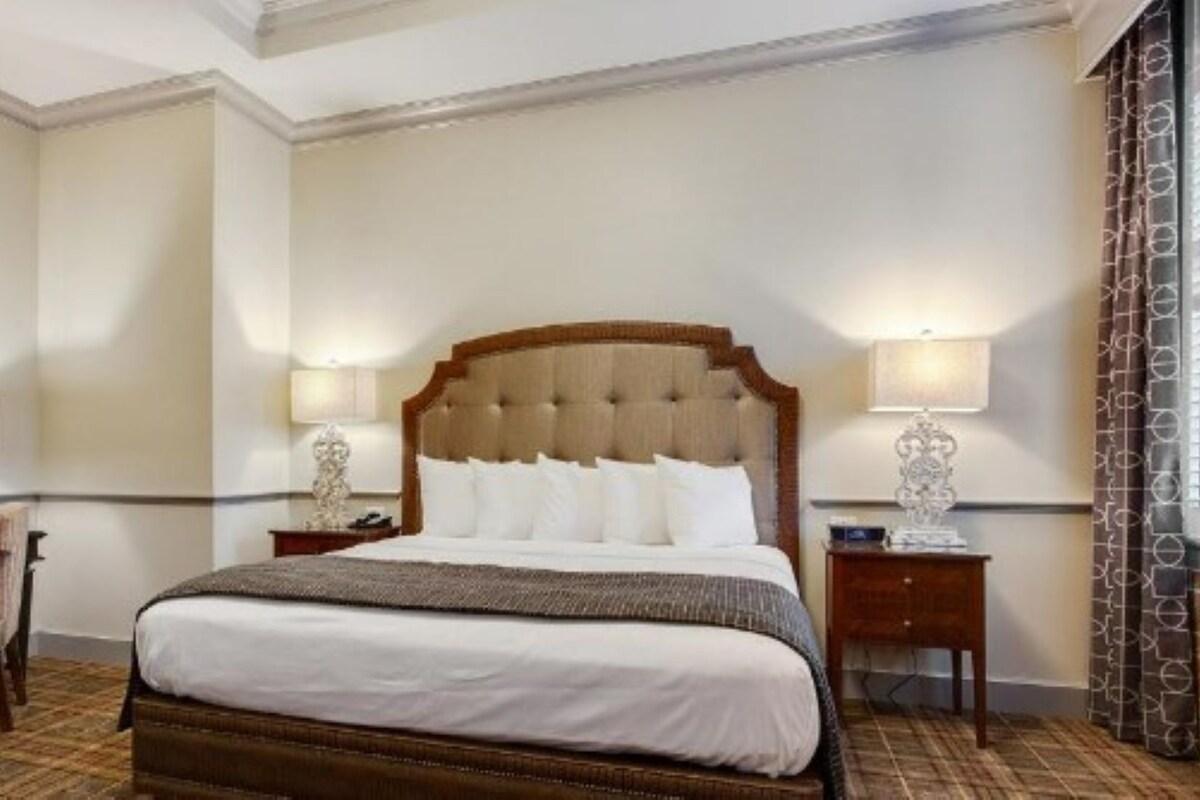 A hotel room with a large bed, two bedside tables, a desk, a chair, and a window with patterned curtains.