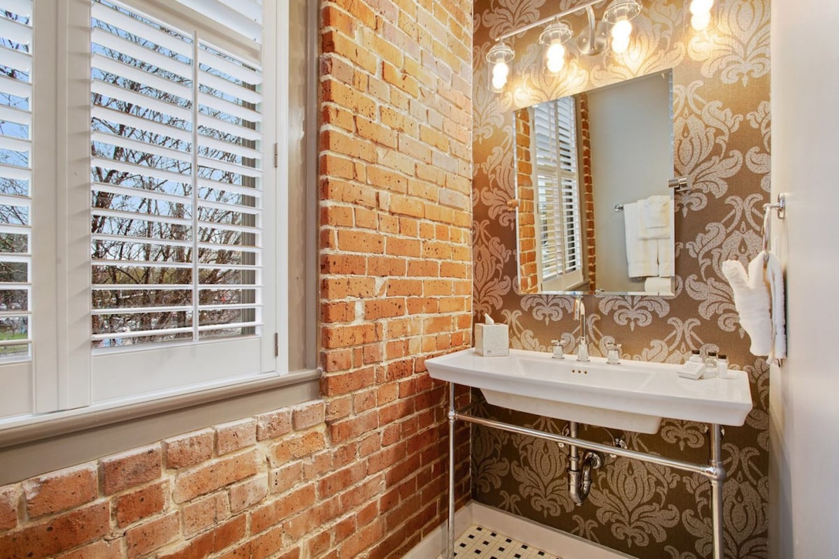 A bathroom with a brick wall, a window with blinds, a sink with a mirror, and a patterned wall.