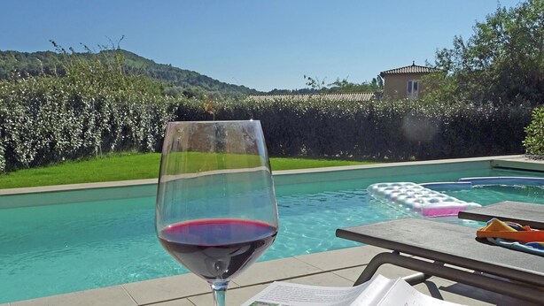 A glass of red wine, a book, and a poolside setting with a clear view of a mountain and a building in the background. - Villa in Malaucene Near Mount Ventoux (Malaucene)