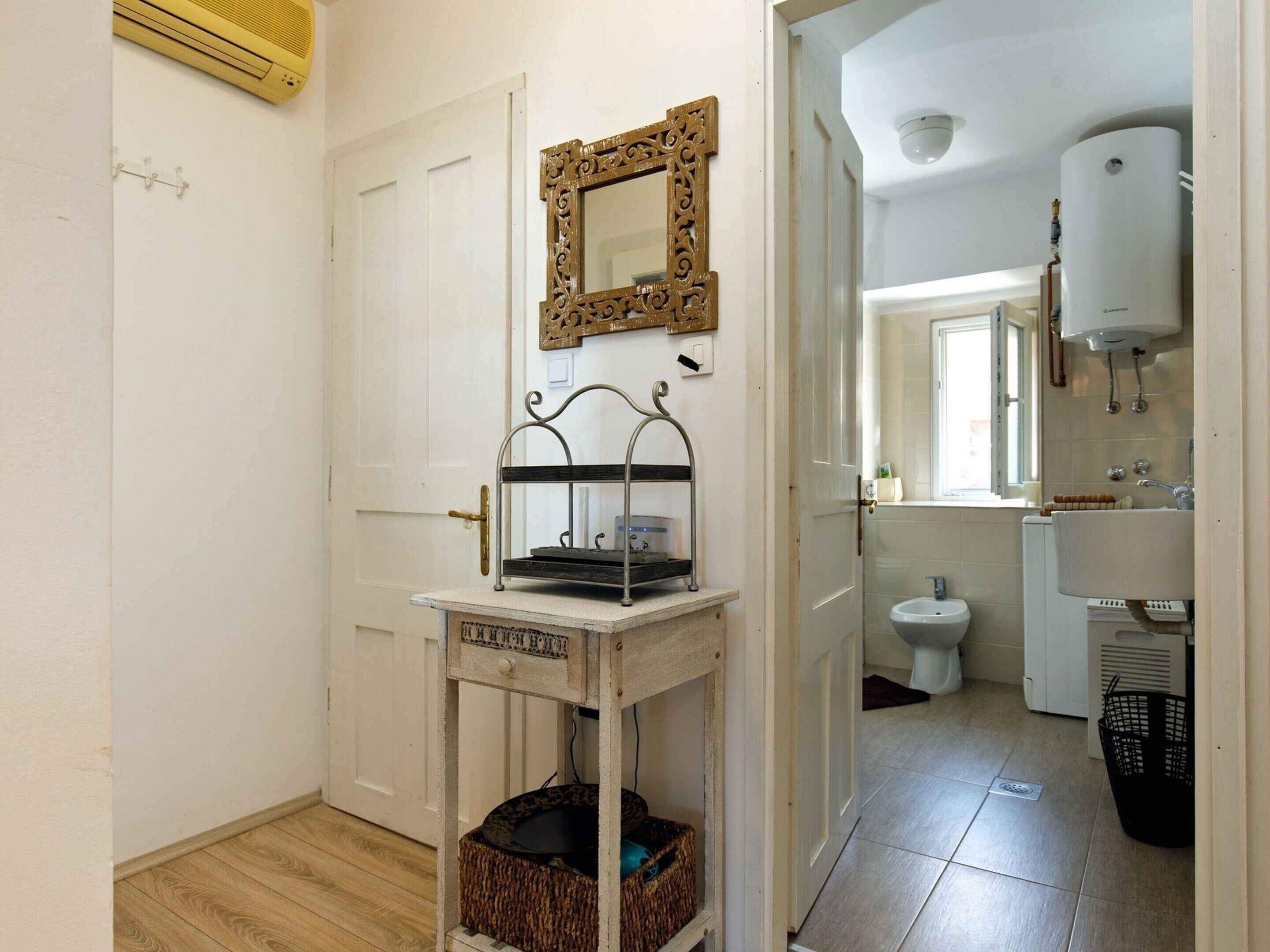 A hallway leading to a bathroom with a white door, a mirror, a small table with a basket, and a radiator.