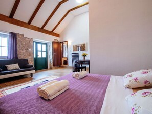 Une chambre avec un lit, un canapé, un bureau et une chaise. On y trouve un mur en pierre et des poutres en bois au plafond.