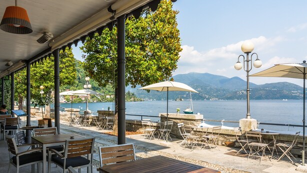 An outdoor café with tables and chairs, umbrellas, and a view of a lake and mountains. - Chalet in Verbania Near Lake Maggiore (Verbania)