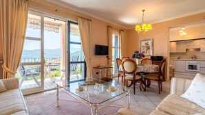 A spacious living room with a glass coffee table, a sofa, a dining area with chairs, a kitchen with cabinets, and a TV mounted on the wall. - Chalet in Verbania Near Lake Maggiore (Verbania)