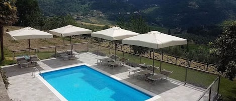 Une piscine rectangulaire à l’eau d’un bleu limpide, entourée d’une terrasse carrelée. Quatre parasols blancs offrent de l’ombre, et plusieurs chaises longues sont disposées autour de la piscine. La terrasse est délimitée par une clôture en métal, et en arrière-plan, on aperçoit un paysage montagneux verdoyant.