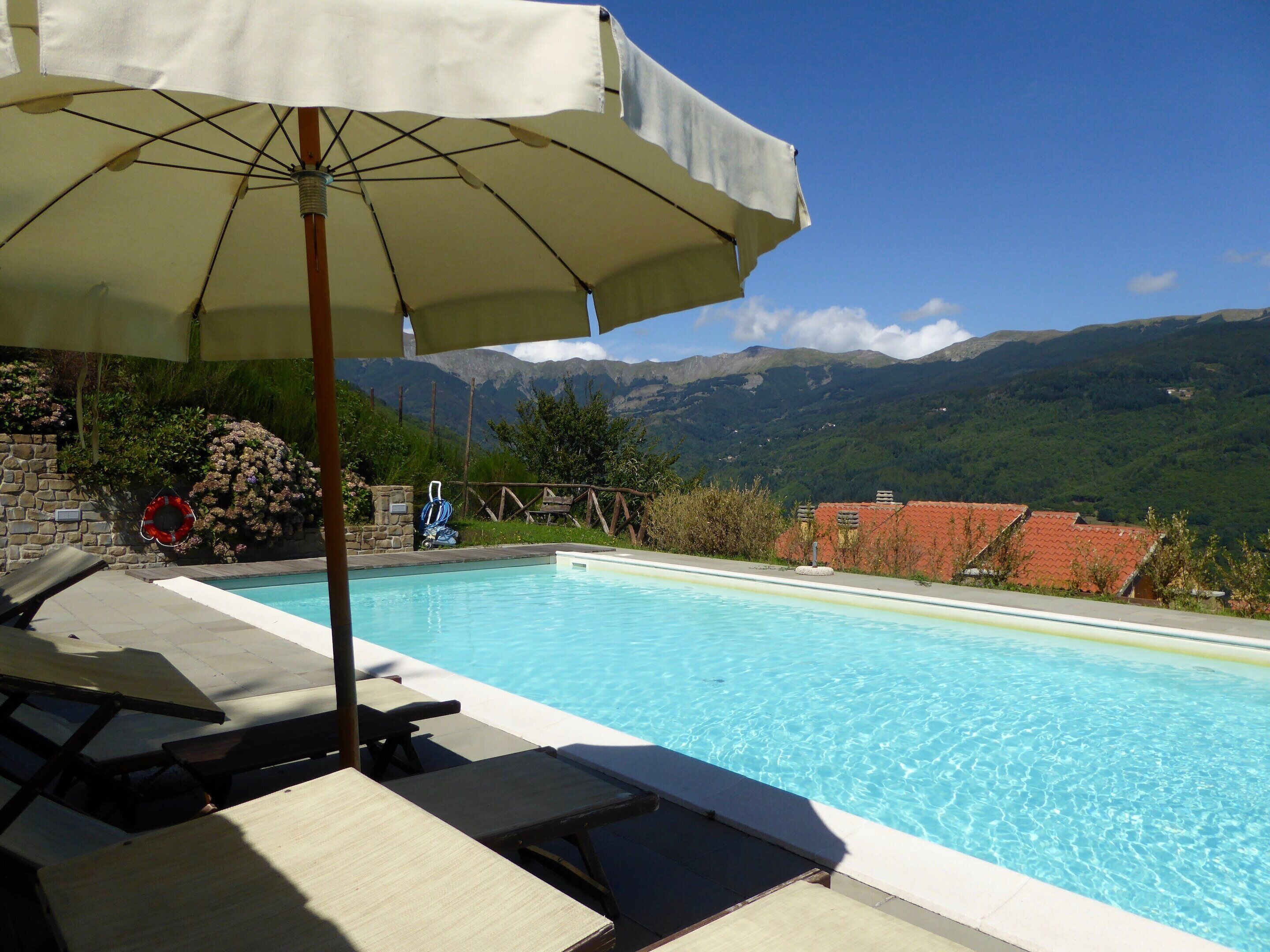 Piscina con sillones de descanso y una sombrilla, rodeada de montañas y follaje.