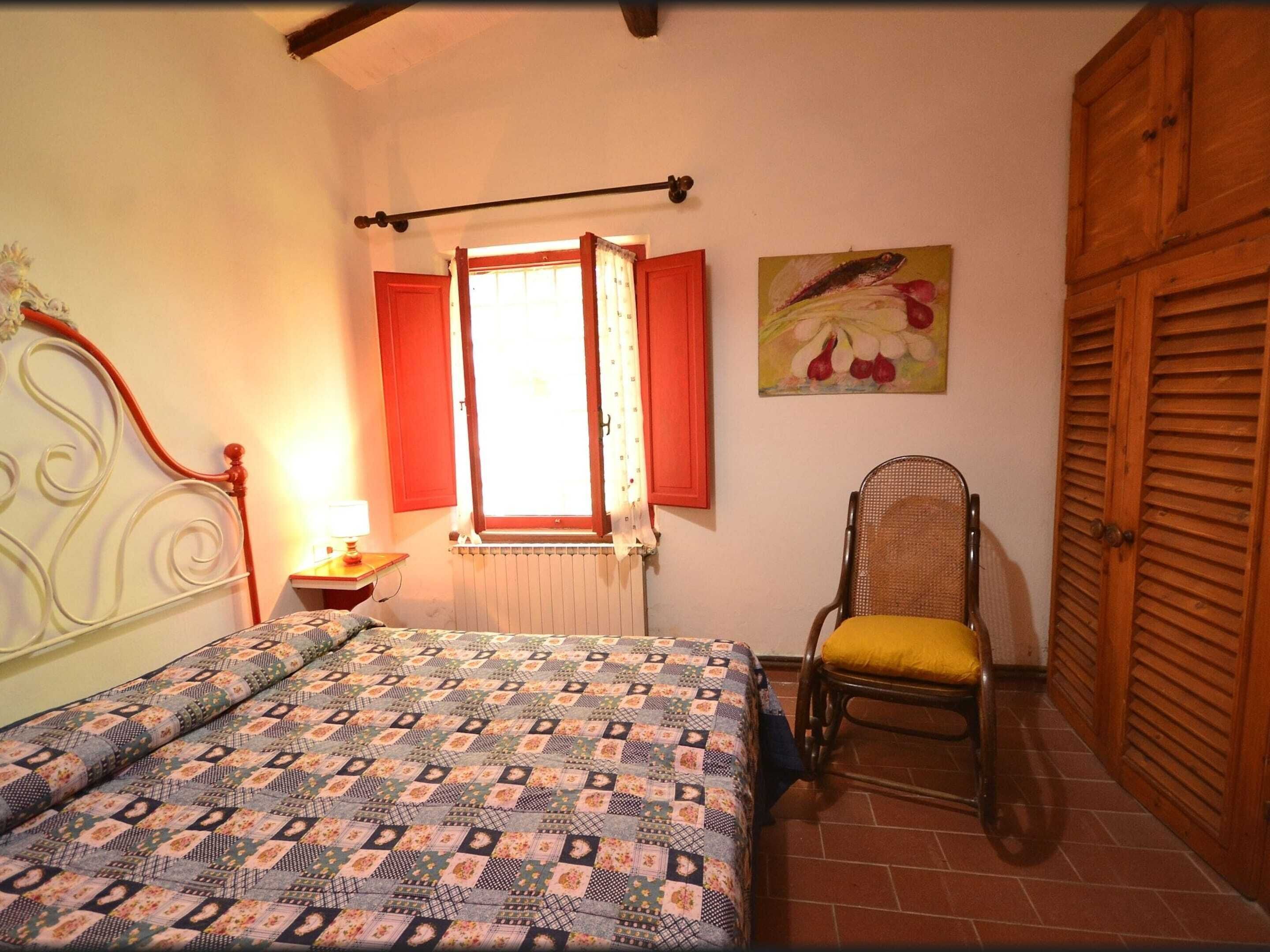 A bedroom with a bed, a chair, a window with red shutters, and a radiator.