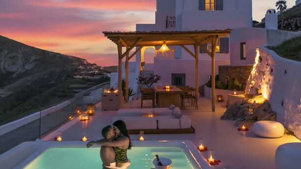 A couple embracing by a pool with a sunset view, a covered seating area, and a white building with a balcony. - Stunning BlueWhite Villa: Luxury Heated Infinity Pool Escape (Πύργος Καλλίστης)