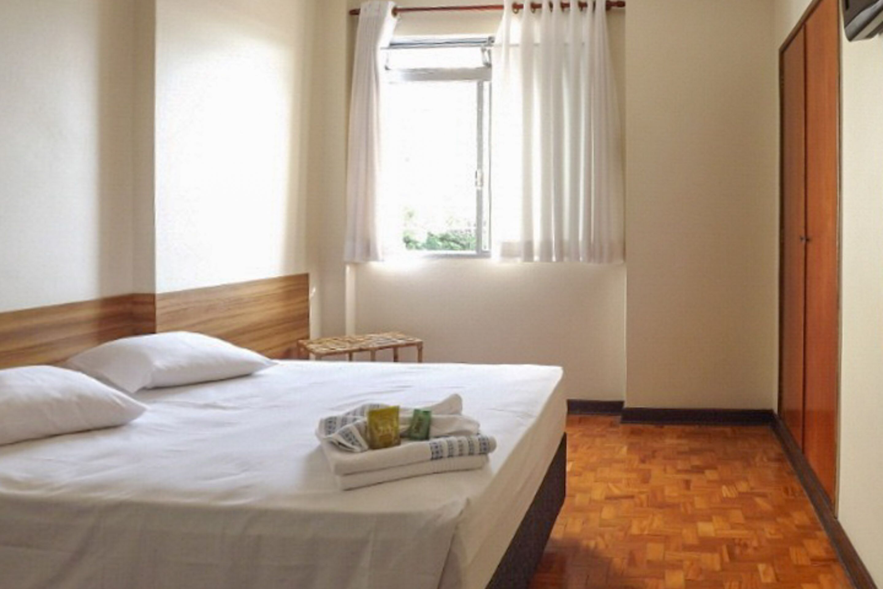 Quarto de hotel com cabeceira de madeira, cama com lençóis brancos, uma mesinha com produtos de higiene, janela com cortinas brancas e piso de madeira.
