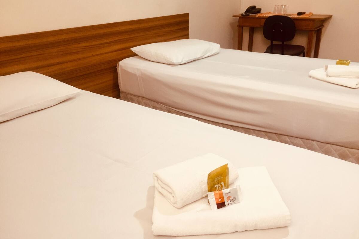 Une chambre d’hôtel avec deux lits simples, une tête de lit en bois, une petite table avec une chaise, et une bouteille de shampoing et d’après-shampoing posée sur les lits.