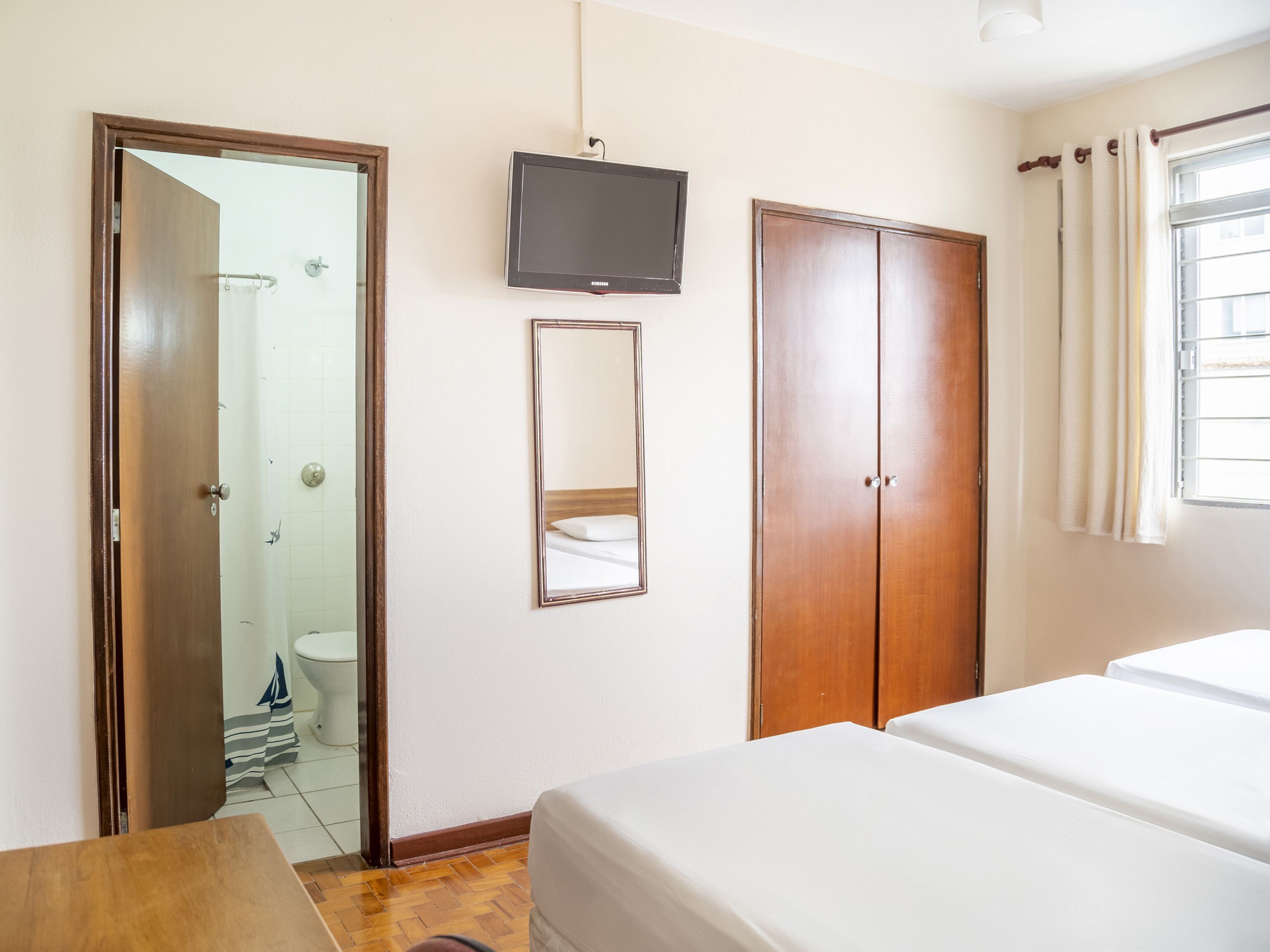 Quarto de hotel com guarda-roupa de madeira, televisão na parede e cama com roupa de cama branca.