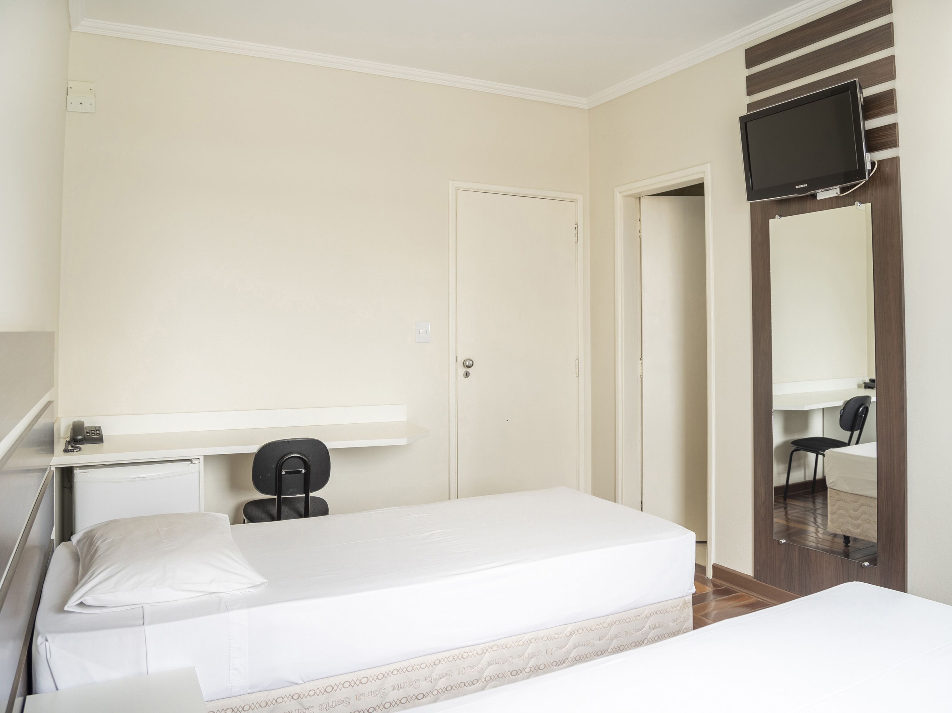A hotel room with a bed, a desk with a chair, a television mounted on the wall, and a door.