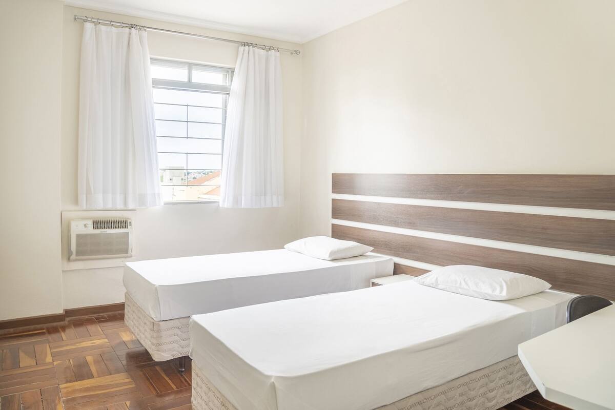 Une chambre d’hôtel avec deux lits simples, une tête de lit en bois, une fenêtre avec des rideaux légers et un climatiseur fixé au mur.