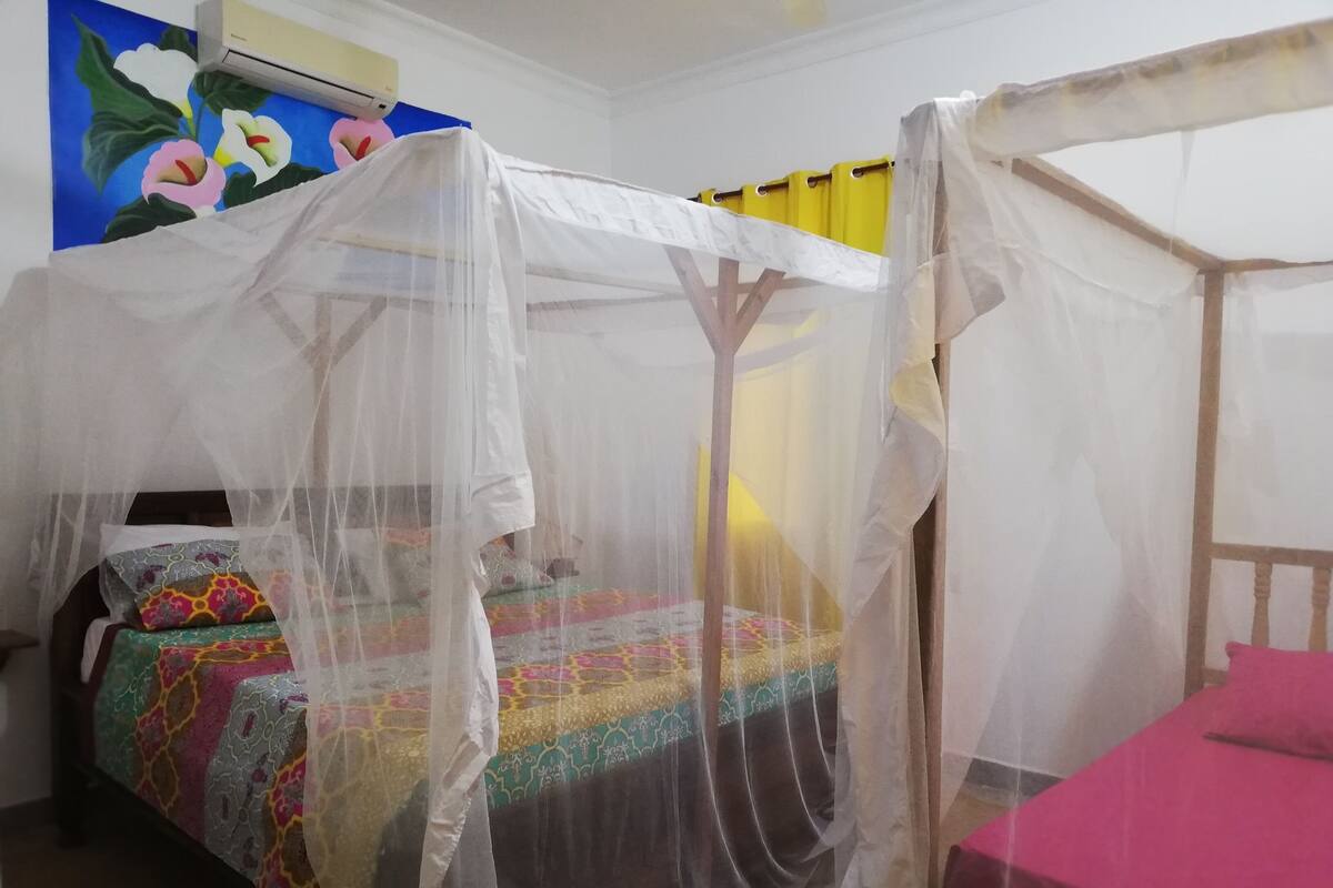 Quarto com cama sob uma rede mosquiteira, mesa de cabeceira, cadeira e parede com pintura floral.