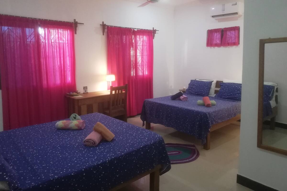 Quarto com duas camas, cada uma com colcha e almofadas azuis, uma escrivaninha com abajur e um ventilador de teto.