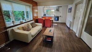 A modern living room with a sofa, a coffee table, and a dining area with red chairs. A kitchen with white cabinets and a dishwasher is visible in the background.
