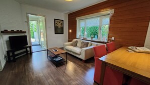 A living room with a sofa, a coffee table, a TV, and a dining table with chairs. There is a window with a view of greenery outside.