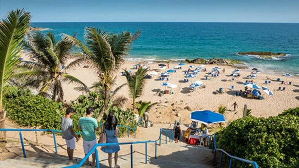 Ein Strand mit Palmen, einem Sandufer und Menschen, die unter Sonnenschirmen entspannen.