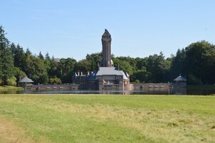 A large, historic building with a prominent tower, situated by a lake with a grassy foreground.