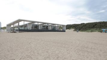 Une maison de plage moderne dotée d’un grand patio ouvert, située sur une plage de sable fin, avec une colline boisée en arrière-plan.
