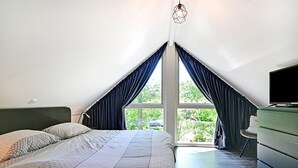 Ein Schlafzimmer mit einem großen Bett, einem Schminktisch und einem Fernseher. Ein Fenster mit Vorhängen bietet Blick auf Bäume.
