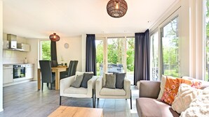 Un salon moderne avec un canapé, des fauteuils et une table basse. On y trouve également un coin repas avec une table et des chaises, et une cuisine à l’arrière-plan.