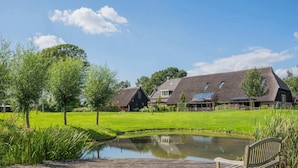 Ein großes Haus mit Teich, umgeben von viel Grün und einer Holzterrasse mit einer Bank.