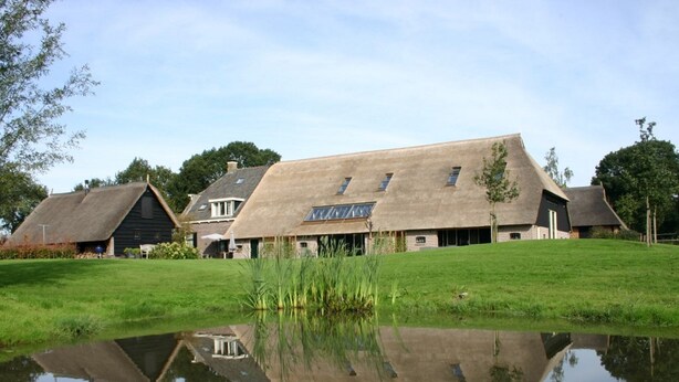 Ein traditionelles Gebäude mit einem strohgedeckten Dach und einem Teich davor, umgeben von viel Grün.