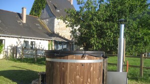 A wooden hot tub with a metal structure beside it, set on a concrete platform in a grassy area with a house in the background. - GITE DE LA BRETAUDERIE 6/8 PEOPLE NEAR ANGERS (RIVES DU LOIR EN ANJOU)