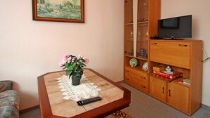 A living room with a wooden cabinet, a television on top, a remote control, a table with a lace tablecloth, and a potted plant. - Hubsches Appartement in Kropelin mit Terrasse (Kroepelin)