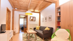 A cozy living room with a wooden ceiling, a grey sofa with yellow cushions, a white TV stand, a small table with a vase, and a green chair. - Cozy Apartment in Rerik near Sea (Rerik)