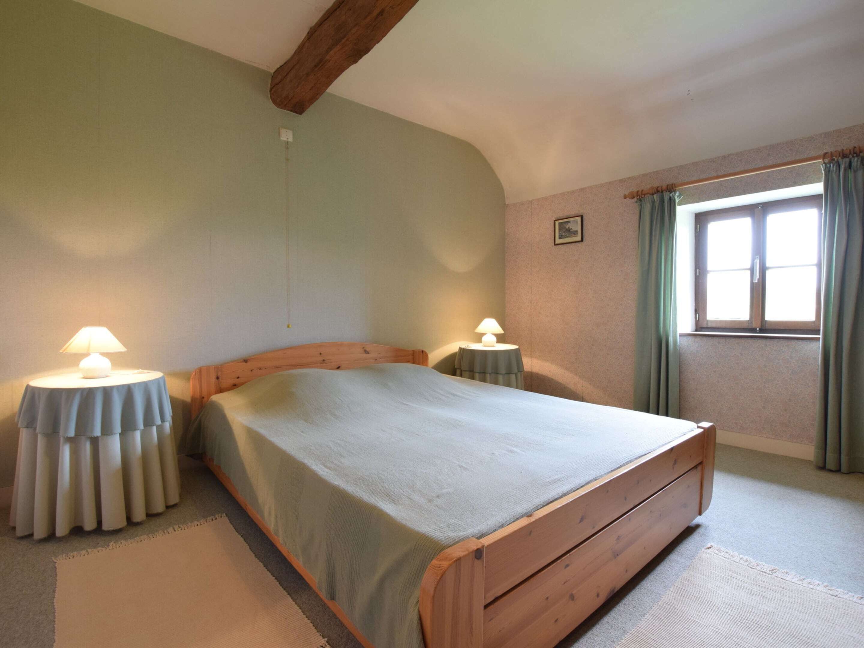 A bedroom with a wooden bed, two bedside tables with lamps, a window with curtains, and a small picture on the wall.